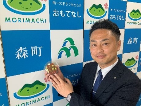 セブン‐イレブン・ジャパンの『北海道森町産トマトとパスタのカプレーゼ仕立て』-商品を持って微笑む岡嶋町長